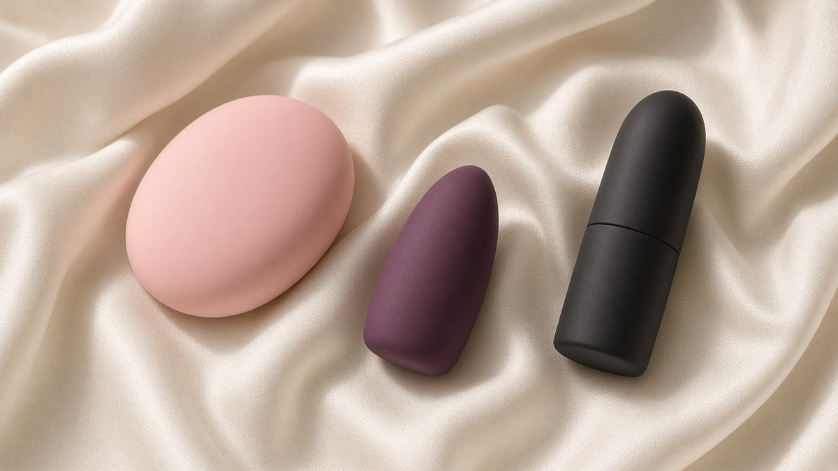 Tres vibradores de clítoris sin marca: uno plano rosa, una bala morada y un pintalabios negro sobre satén marfil