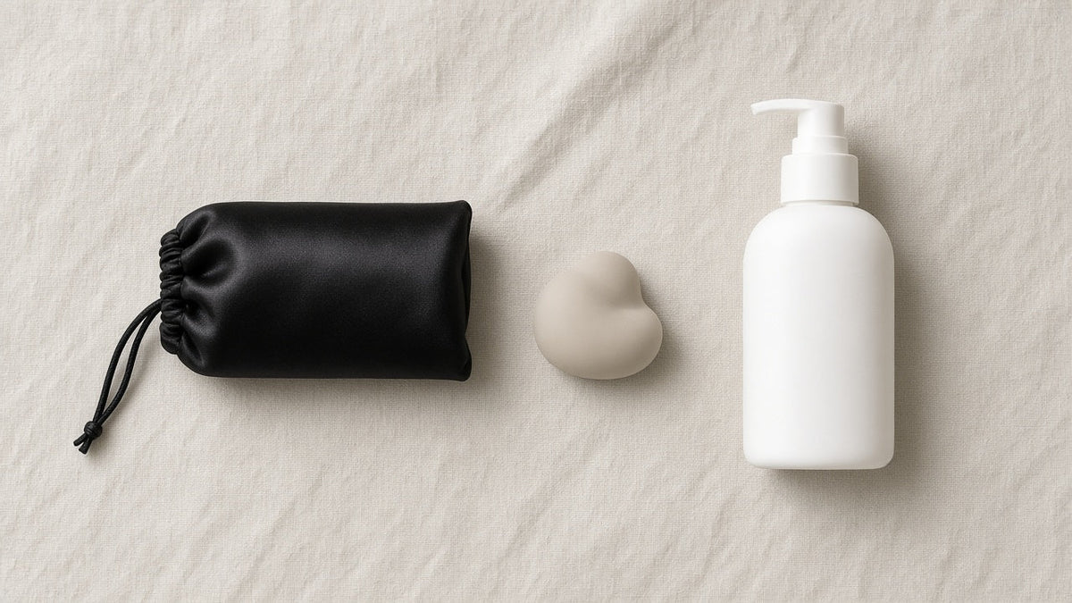 Bolsa de satén negra, pieza de silicona neutra y frasco dosificador sobre lino, para la guía sobre fundas de pene